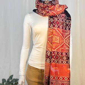 May include: A colorful scarf with a geometric pattern and fringe. The scarf features shades of red, orange, and purple. The design includes geometric shapes and images of animals. The scarf is draped over a white long-sleeve shirt.