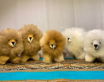 Op de afbeelding: Vijf pluche leeuwen speelgoed gemaakt van alpaca wol. De leeuwen zijn in een rij op een kleurrijk tapijt gerangschikt. De leeuwen zijn bruin, beige en wit.