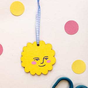 May include: A yellow scalloped wooden sun ornament featuring a hand-painted face with dark blue eyes, eyebrows, and a smile, along with pink cheeks. The sun is suspended by a light blue and white gingham ribbon loop for hanging purposes.