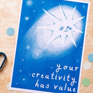 以下が含まれることがあります： 笑顔の太陽のデザインと白い文字「Your creativity has value」が特徴の青いアートプリント。木製の表面に散らばった紙吹雪が添えられています。