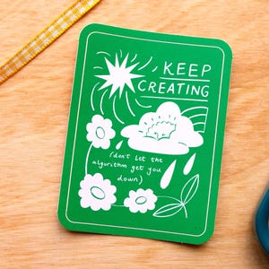 May include: A green rectangular sticker with white illustrations and text. The text reads "KEEP CREATING" above a cloud and flower design. Below the design is the text "(don't let the algorithm get you down)".