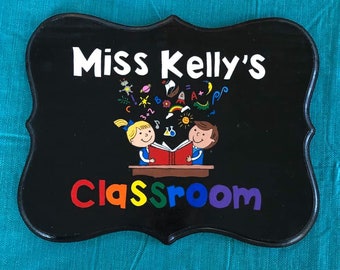 May include: Black wooden sign with a white inscription that reads "Miss Kelly's Classroom". The sign is decorated with colourful illustrations of children reading a book and other school-related objects.