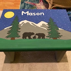 May include: A green wooden step stool with a blue, white, and gray painted scene of a mountain range, two pine trees, a full moon, and a family of bears. The name "Mason" is written in white letters on the top of the stool.