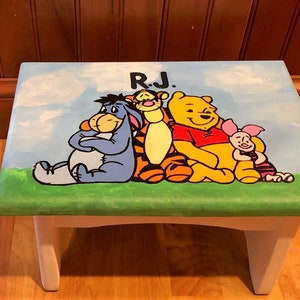 May include: Hand-painted wooden step stool featuring Winnie the Pooh and friends. The stool has a blue top with a green border, white legs, and the initials "R.J." painted on top. The characters include Eeyore, Tigger, Pooh, and Piglet.