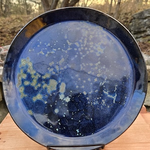 Peut inclure: Une assiette en céramique faite à la main avec un glaçage bleu profond et des accents mouchetés. L'assiette a un large bord et est présentée sur un support en métal noir. Le glaçage présente des variations de bleu et des touches de vert.