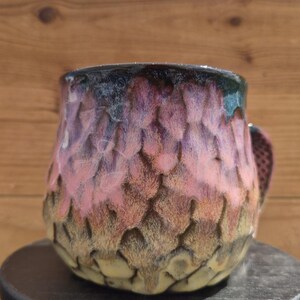 May include: A ceramic mug featuring a textured, scale-like pattern across its surface. The glaze transitions from a deep, dark rim down through shades of vibrant pink and purple, settling into a muted, earthy yellow-brown tone at the base.