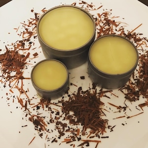 May include: Three small round containers of yellow balm, with a brown, shredded material scattered around them on a white surface.