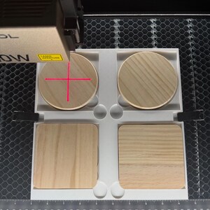 May include: A laser engraving machine in operation, focused on four wooden coasters. Two round and two square coasters are arranged in a white tray. The laser's red crosshairs are visible on one of the round coasters. The machine has a gold and black finish.