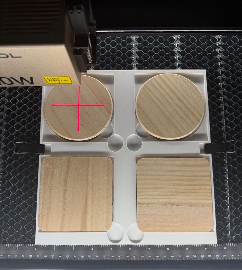 May include: A laser engraving machine in operation, focused on four wooden coasters. Two round and two square coasters are arranged in a white tray. The laser's red crosshairs are visible on one of the round coasters. The machine has a gold and black finish.
