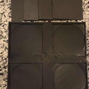 May include: A set of dark gray slate coasters and a black plastic coaster holder. The coasters are rectangular and square, while the holder has four circular spaces for the coasters. The holder has a unique design.