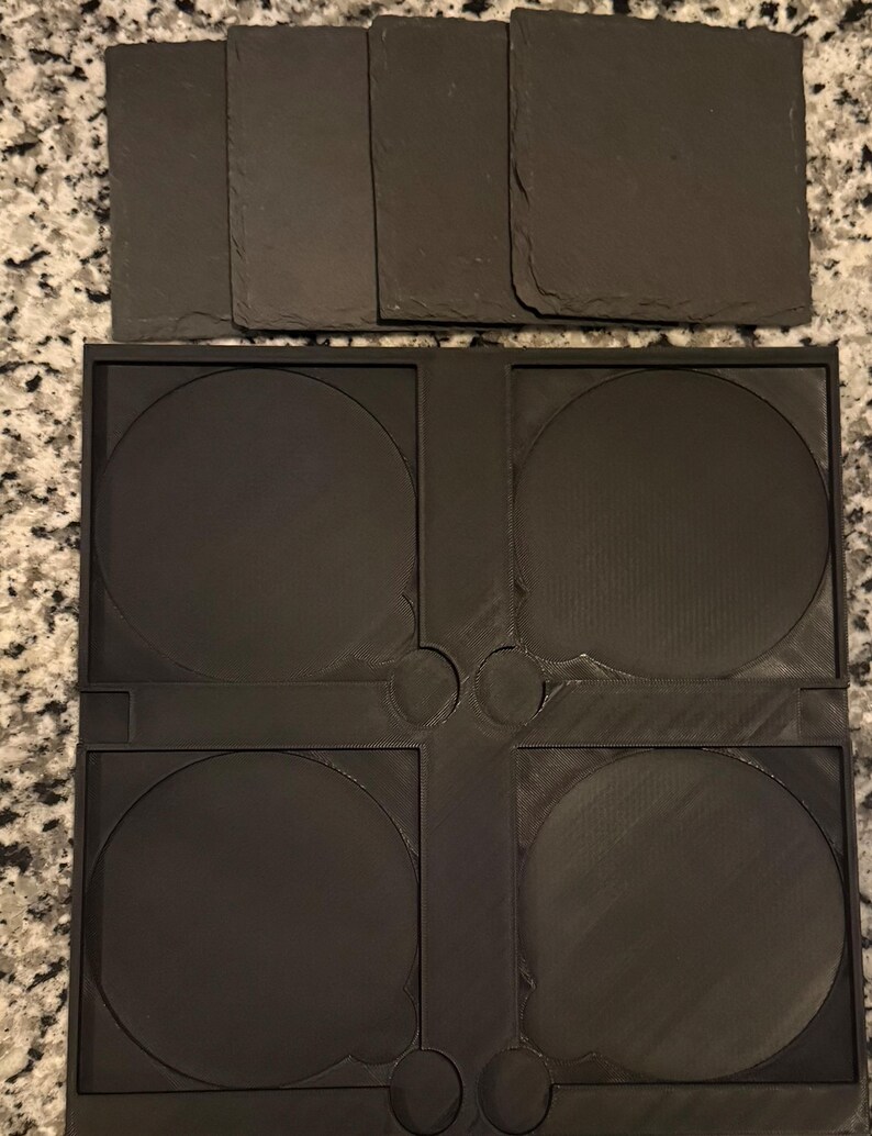 May include: A set of dark gray slate coasters and a black plastic coaster holder. The coasters are rectangular and square, while the holder has four circular spaces for the coasters. The holder has a unique design.