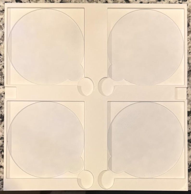 May include: A white, square, four-sectioned tray with rounded cutouts. Each section features a circular indentation with a small, rounded tab. The tray is designed to hold and organize small, round objects.