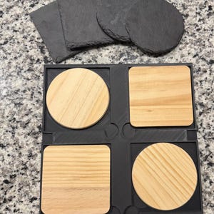May include: A set of coasters in a variety of shapes and materials. The image shows four square and round wooden coasters in a black holder, along with several slate coasters. The wooden coasters have a light brown color and visible wood grain.