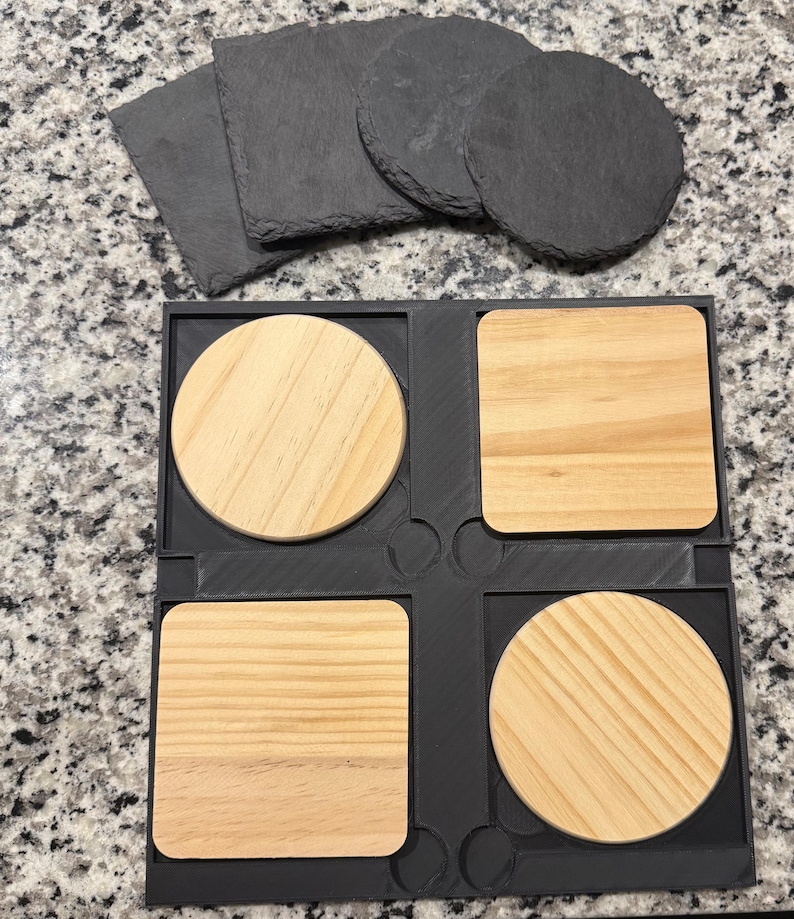 May include: A set of coasters in a variety of shapes and materials. The image shows four square and round wooden coasters in a black holder, along with several slate coasters. The wooden coasters have a light brown color and visible wood grain.