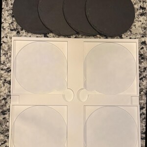 May include: A set of four round, dark gray slate coasters arranged above a white plastic tray with four circular indentations. The coasters have a natural, textured surface, perfect for protecting surfaces from drink condensation.