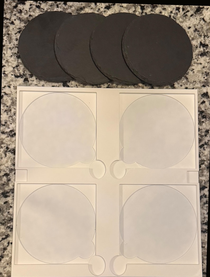 May include: A set of four round, dark gray slate coasters arranged above a white plastic tray with four circular indentations. The coasters have a natural, textured surface, perfect for protecting surfaces from drink condensation.