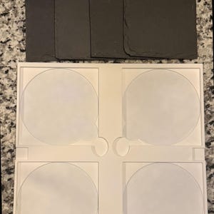 May include: A set of four dark gray slate coasters with a natural, textured surface. Below, a white plastic holder with four circular cutouts designed to hold the coasters. The coasters are approximately 4 inches square.