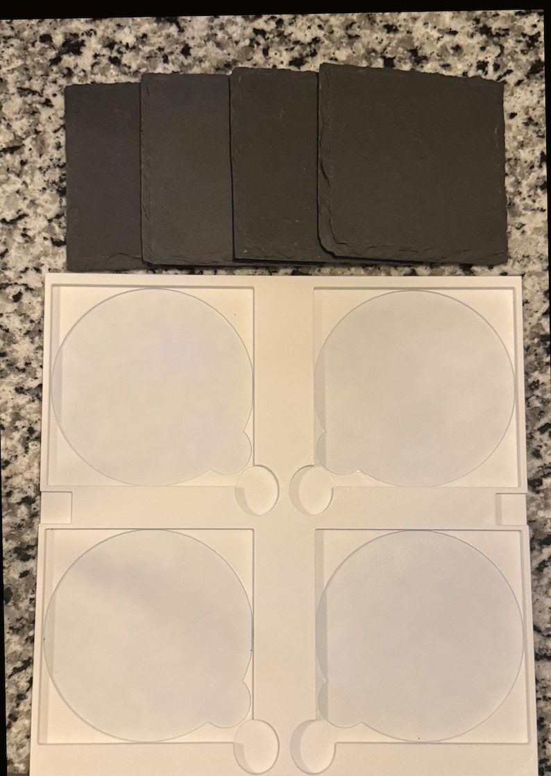 May include: A set of four dark gray slate coasters with a natural, textured surface. Below, a white plastic holder with four circular cutouts designed to hold the coasters. The coasters are approximately 4 inches square.