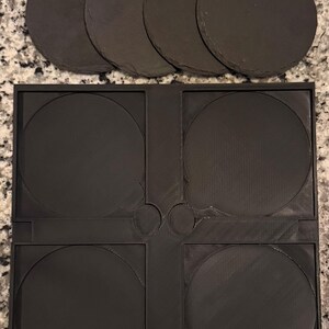 May include: A set of five round, dark gray slate coasters and a black plastic coaster holder. The coasters are made of natural stone and have a rough texture. The holder has four circular indentations to hold the coasters.
