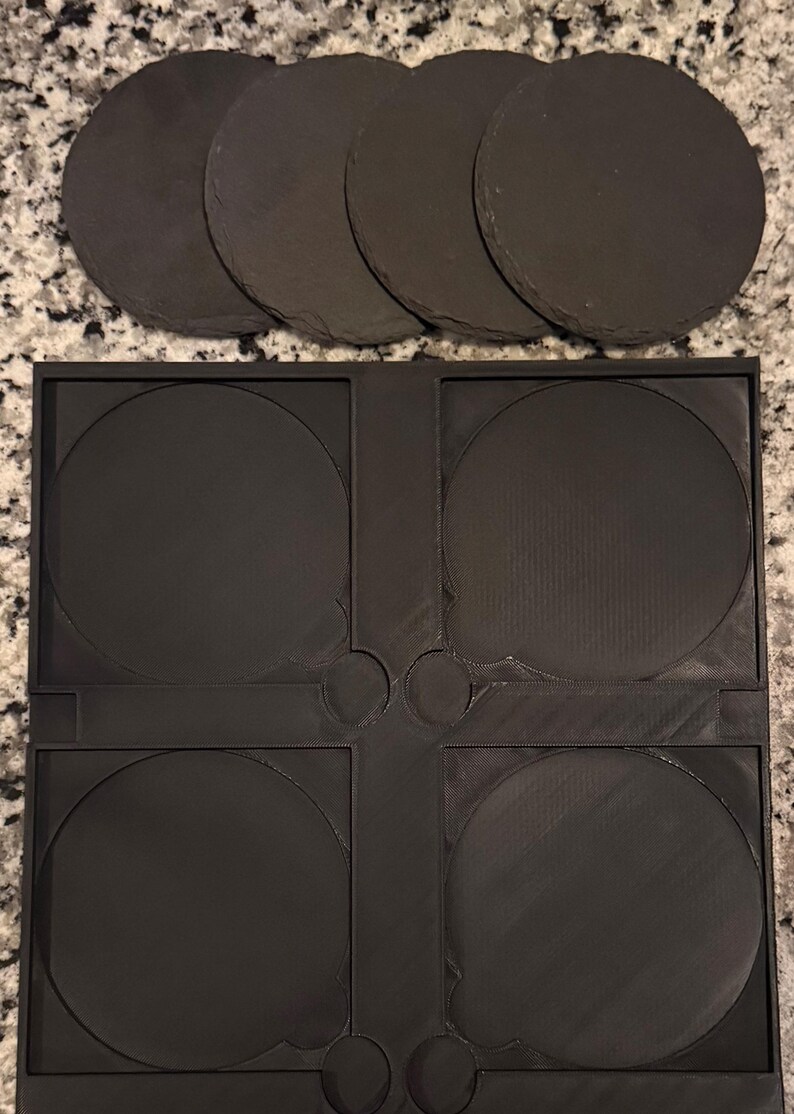 May include: A set of five round, dark gray slate coasters and a black plastic coaster holder. The coasters are made of natural stone and have a rough texture. The holder has four circular indentations to hold the coasters.
