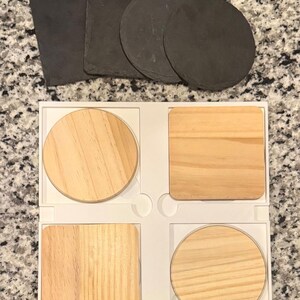 May include: A set of coasters in various shapes and materials. There are four wooden coasters in a white tray, two square and two round. Above them are three round and two square slate coasters. The coasters are designed to protect surfaces from heat and moisture.