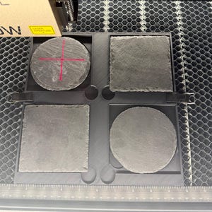 May include: Four slate coasters in a tray, two square and two round. The coasters are a dark gray color and have a rough texture. The tray is black and sits on a metal surface. A laser tool is positioned above the coasters.