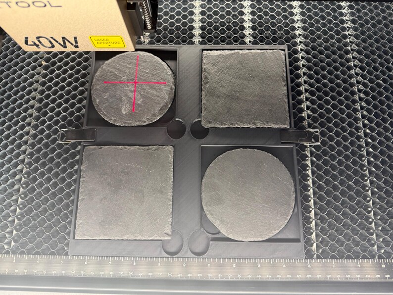 May include: Four slate coasters in a tray, two square and two round. The coasters are a dark gray color and have a rough texture. The tray is black and sits on a metal surface. A laser tool is positioned above the coasters.