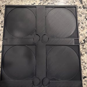 May include: A black plastic CD or DVD storage case with four circular compartments. The case has a rectangular shape with rounded corners and a textured surface. The compartments are designed to hold discs securely.