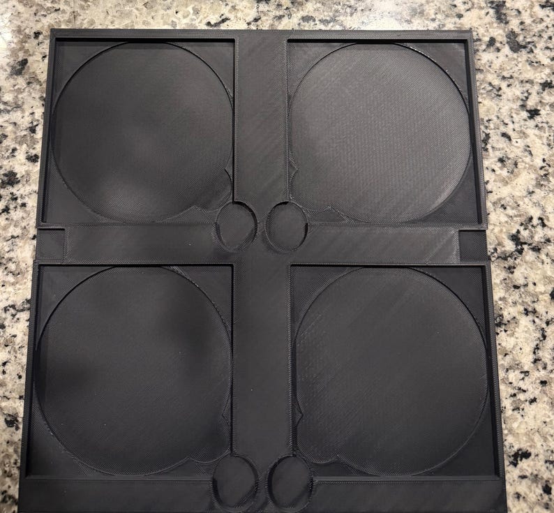 May include: A black plastic CD or DVD storage case with four circular compartments. The case has a rectangular shape with rounded corners and a textured surface. The compartments are designed to hold discs securely.