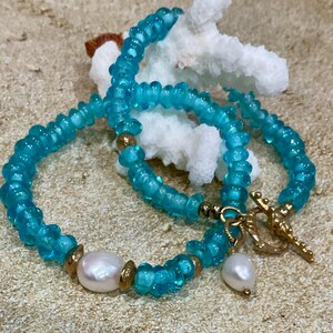 Puede incluir: Un collar de cuentas turquesas con cierre y detalles dorados. El collar presenta una perla blanca grande y un colgante de perla más pequeño en forma de lágrima. Las cuentas son de color azul claro translúcido.