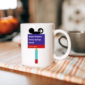 Op de afbeelding: Witte keramische mok met een kleurrijke afbeelding van een Mickey Mouse-hoofd en een verkeersbord met de tekst "Magic Kingdom, Disney Springs, EPCOT, Rechtsaf".