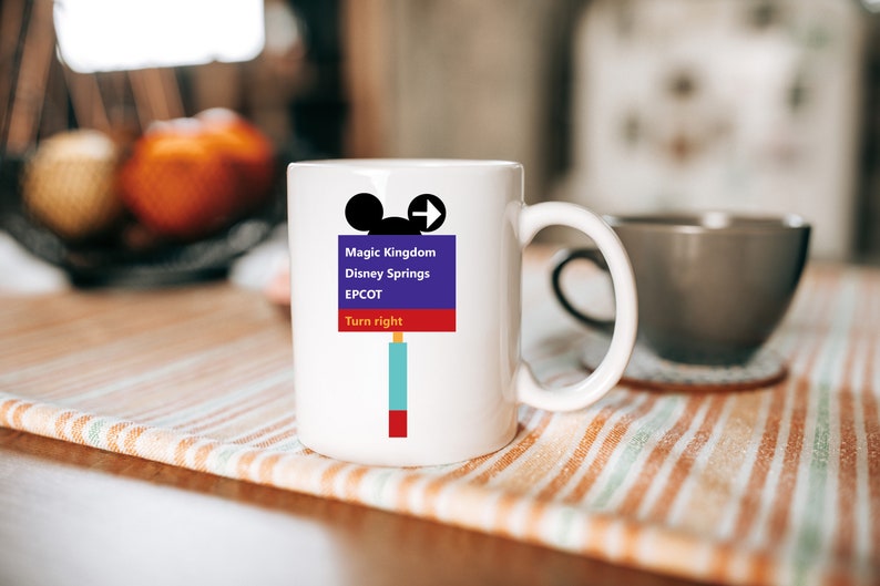 Op de afbeelding: Witte keramische mok met een kleurrijke afbeelding van een Mickey Mouse-hoofd en een verkeersbord met de tekst "Magic Kingdom, Disney Springs, EPCOT, Rechtsaf".