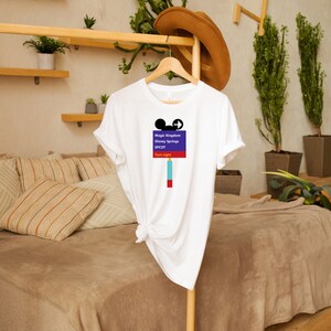Op de afbeelding: Wit T-shirt met een paars en blauw bord waarop staat "Magic Kingdom, Disney Springs, EPCOT, Rechtsaf slaan". Het bord heeft een Mickey Mouse-hoofd en een pijl die naar rechts wijst.