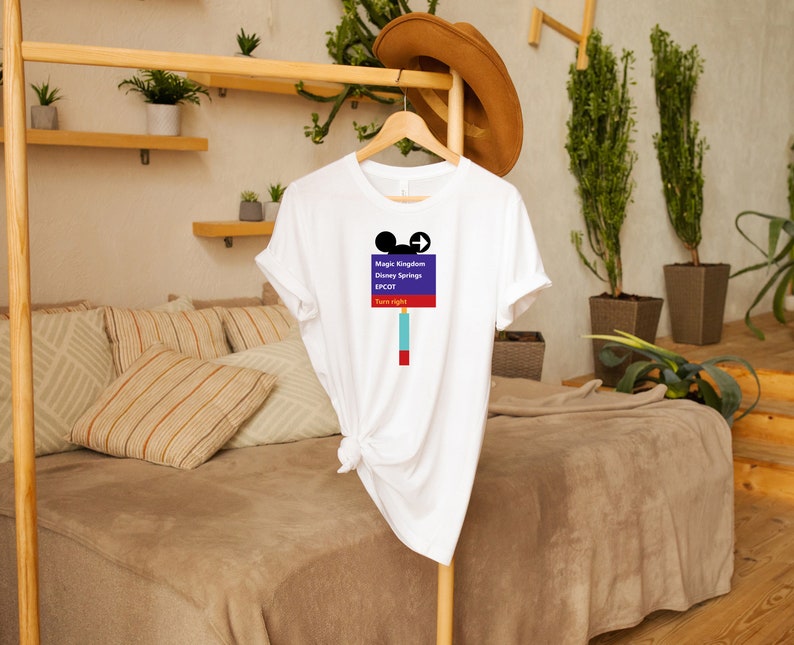 Op de afbeelding: Wit T-shirt met een paars en blauw bord waarop staat "Magic Kingdom, Disney Springs, EPCOT, Rechtsaf slaan". Het bord heeft een Mickey Mouse-hoofd en een pijl die naar rechts wijst.