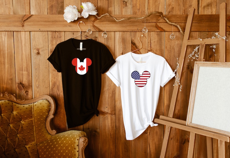 May include: Two t-shirts with a Mickey Mouse head design. The left shirt is black with a Canadian flag inside the Mickey Mouse head. The right shirt is white with an American flag inside the Mickey Mouse head.