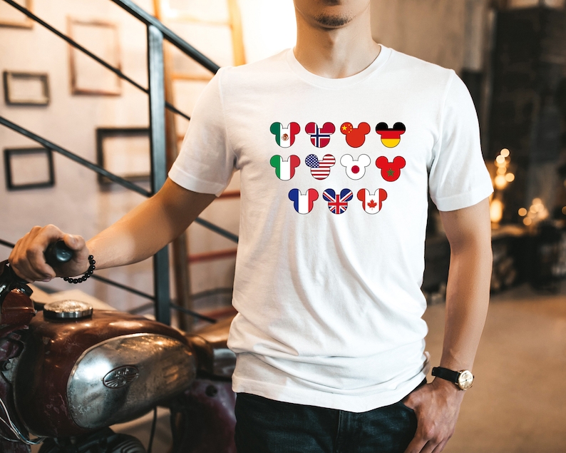 May include: White t-shirt with a design of Mickey Mouse ears with different country flags inside each ear. The flags include Mexico, Norway, China, Germany, Italy, United States, Japan, Morocco, France, United Kingdom, and Canada.