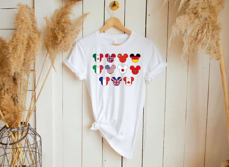 May include: White t-shirt with a design of Mickey Mouse heads with different country flags on them. The flags include Mexico, Norway, China, Germany, Italy, USA, Japan, Morocco, France, UK, and Canada.