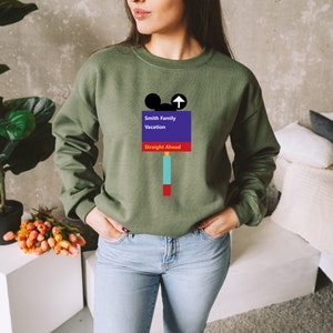 Op de afbeelding: Een groen sweatshirt met een afbeelding van een Mickey Mouse-hoofd en een bord met de tekst "Smith Family Vacation Straight Ahead".