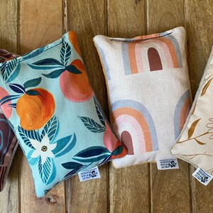 May include: Four small, rectangular fabric pillows with different patterns. The pillows are made of a soft, natural fabric and have a subtle sheen. The patterns include a peach and leaf design, a rainbow design, a floral design, and a geometric design. Each pillow has a small label sewn on the bottom corner that says "Sentient Supply Co."