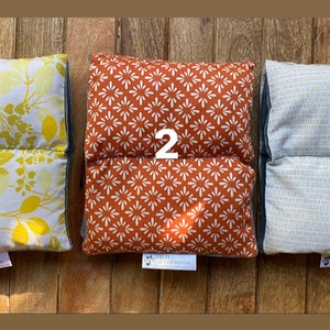 May include: Three rectangular pillows with different patterns. The first pillow has a white background with yellow leaves, the second pillow has a red background with white star-shaped patterns, and the third pillow has a white background with a light gray grid pattern. Each pillow has a small tag with the text "The Shepherd's Hand" on it.