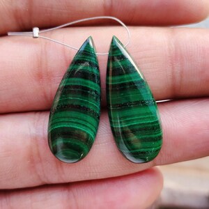 May include: Two green malachite teardrop shaped gemstone beads with black veining. The beads are strung on a silver wire.