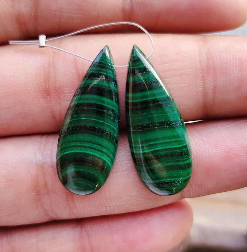 May include: Two green malachite teardrop shaped gemstone beads with black veining. The beads are strung on a silver wire.