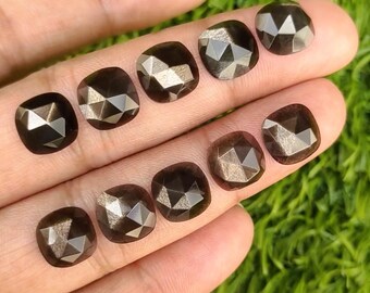 Pode incluir: Uma coleção de dez pedras preciosas facetadas de corte quadrado em tons variados de castanho. As pedras são exibidas em uma mão contra um fundo de folhagem verde. Cada pedra tem um padrão geométrico único.