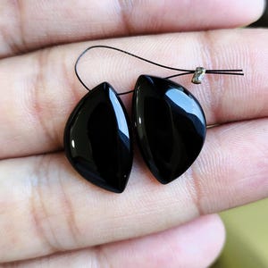 May include: Two polished, black, teardrop-shaped gemstones with smooth, reflective surfaces. Each stone features a drilled hole at the top, and they are connected by a thin, dark string with a small metallic crimp bead visible near the top edge.