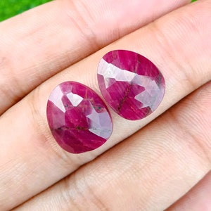 Könnte beinhalten: Zwei rote Rubin-Edelsteine mit facettiertem Schliff. Die Edelsteine sind oval geformt und haben eine tiefrote Farbe.