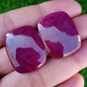 Puede incluir: Dos gemas facetadas, de talla cojín, en un tono magenta intenso. Las gemas se sostienen en una mano sobre un fondo verde borroso. Las piedras tienen una superficie pulida y reflectante, mostrando juegos de luz internos.