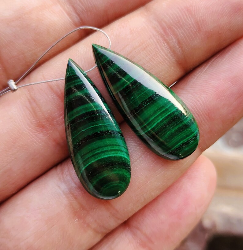 May include: A pair of green malachite teardrop-shaped gemstone pendants with a silver wire loop for hanging.