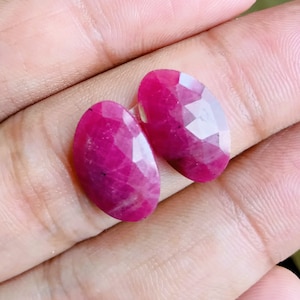 May include: Two faceted, oval-shaped ruby gemstones. The rubies are a deep, vibrant pink-red color with a polished, reflective surface. The gemstones are displayed against a neutral background.