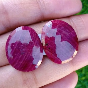 Op de afbeelding: Twee ovaalvormige, gefacetteerde robijnen met een dieprode kleur. De edelstenen worden in een hand gehouden, waarbij hun gepolijste oppervlakken en interne patronen worden getoond. De robijnen hebben een lichtroze tint en zijn geplaatst tegen een vage groene achtergrond.