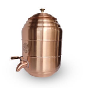 May include: A cylindrical copper water dispenser with a brushed metallic finish, featuring a domed lid with a small knob handle and a spigot at the base. The side is embossed with a circular logo containing the text American Ayurveda.